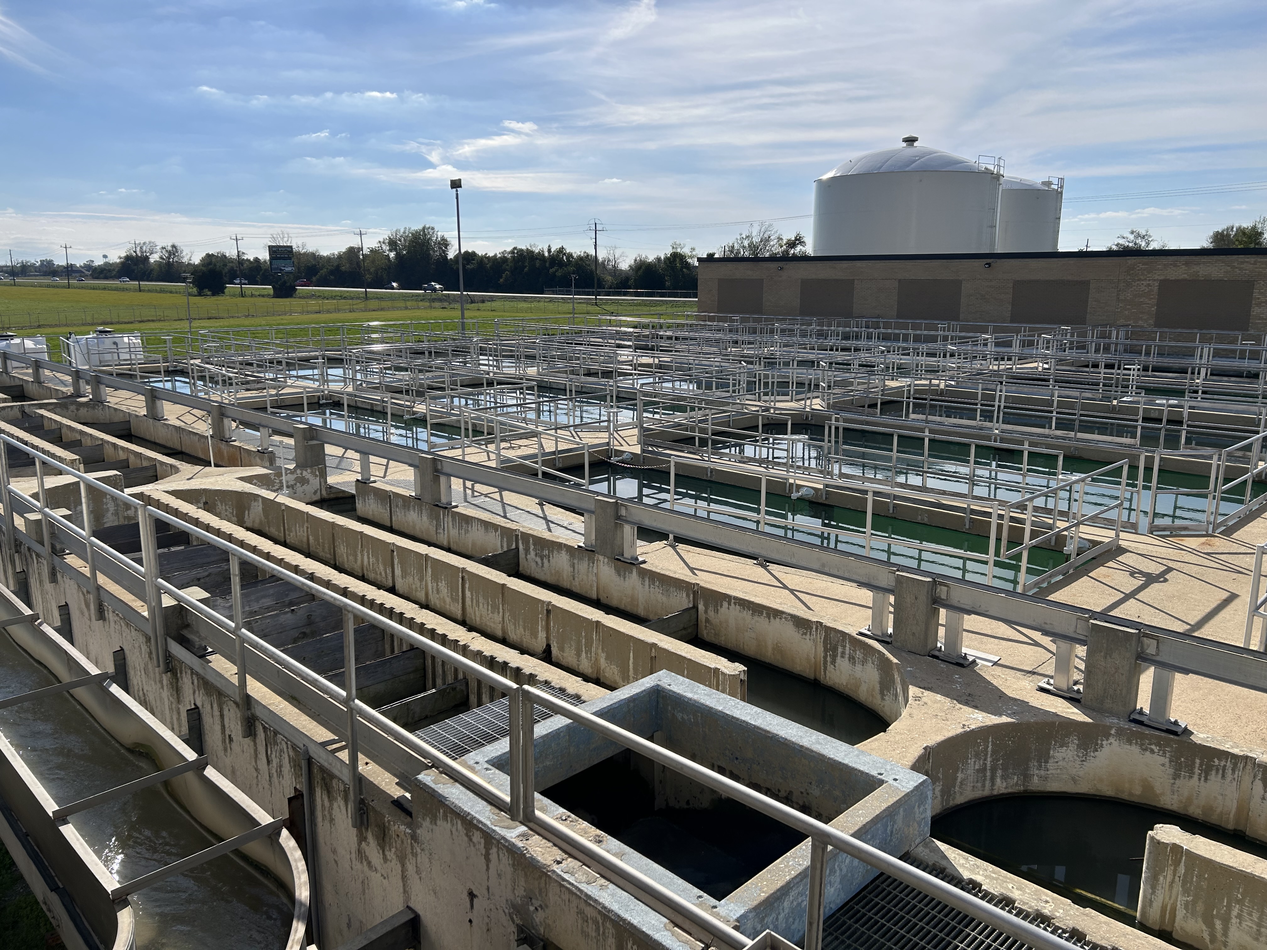 In this photo of a drinking water cleaning facility, pipes and machinery criss cross in front of water pools. 